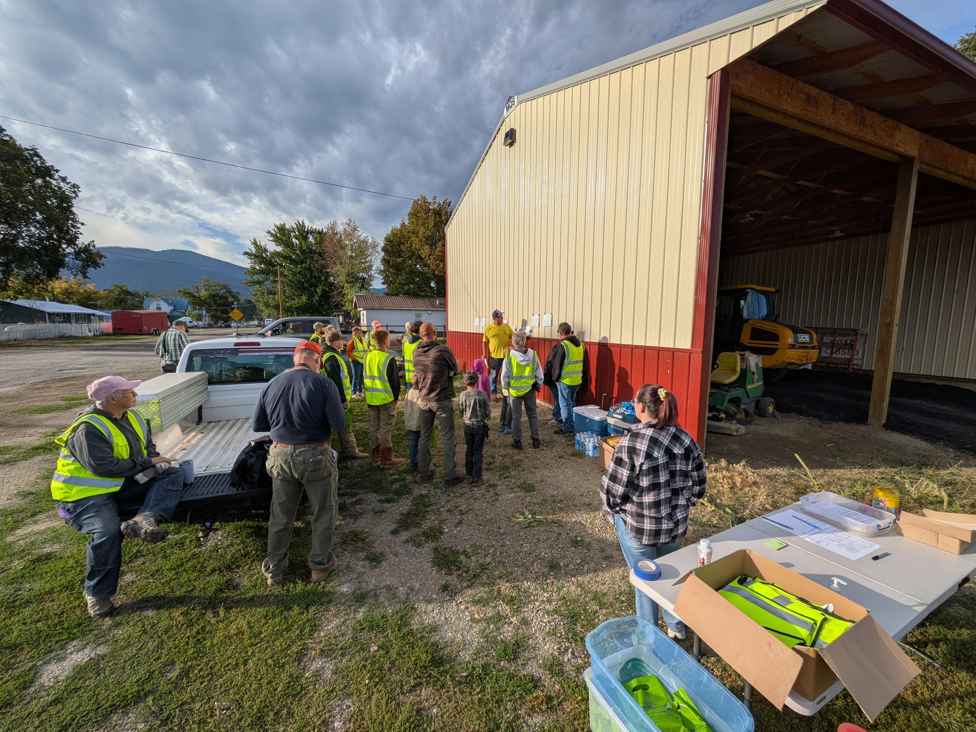 Volunteers in safety vests gathered by a red and tan pole barn for the morning briefing